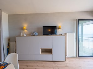 A modern living room with a TV on a cabinet, two lamps, and a large window. - Modern studio with sea view and garage (Middelkerke)