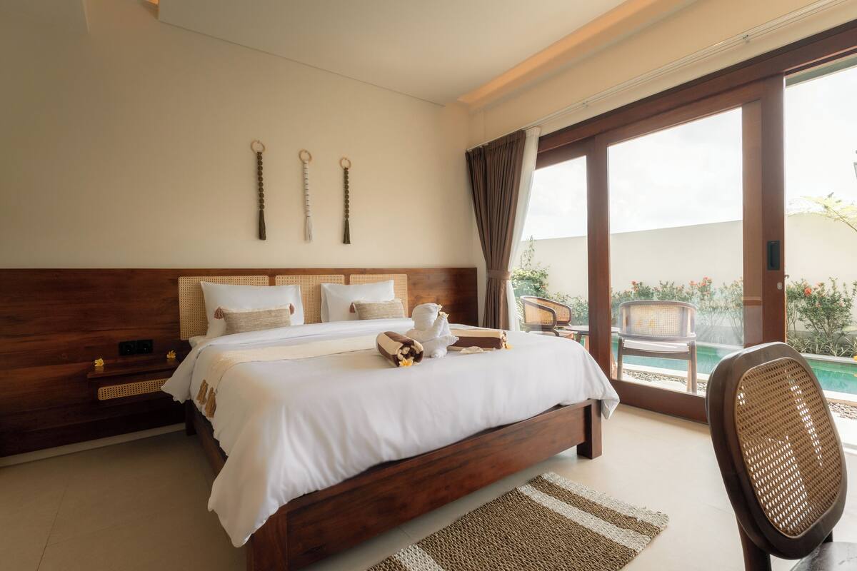 A bedroom with a large bed, wooden headboard, and a view of a swimming pool through a glass door.