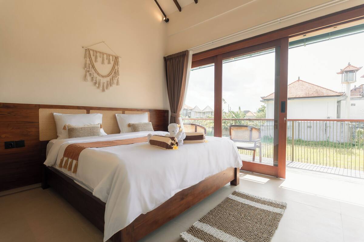 A spacious bedroom with a large bed, a wooden headboard, and a view through a sliding glass door to the outside.