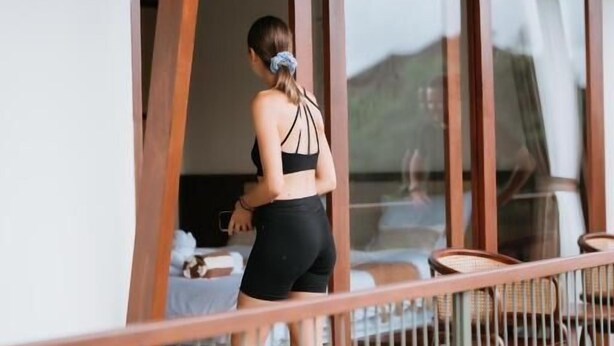 Una persona con top deportivo y pantalones cortos en un balcón con barandas de madera y un ventanal que da a una habitación con cama y mesa.
