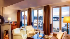 A cozy living room with a fireplace, comfortable seating, and large windows offering a view of the outdoors. - Sunday Resort Hafendorf Suite (Rheinsberg)
