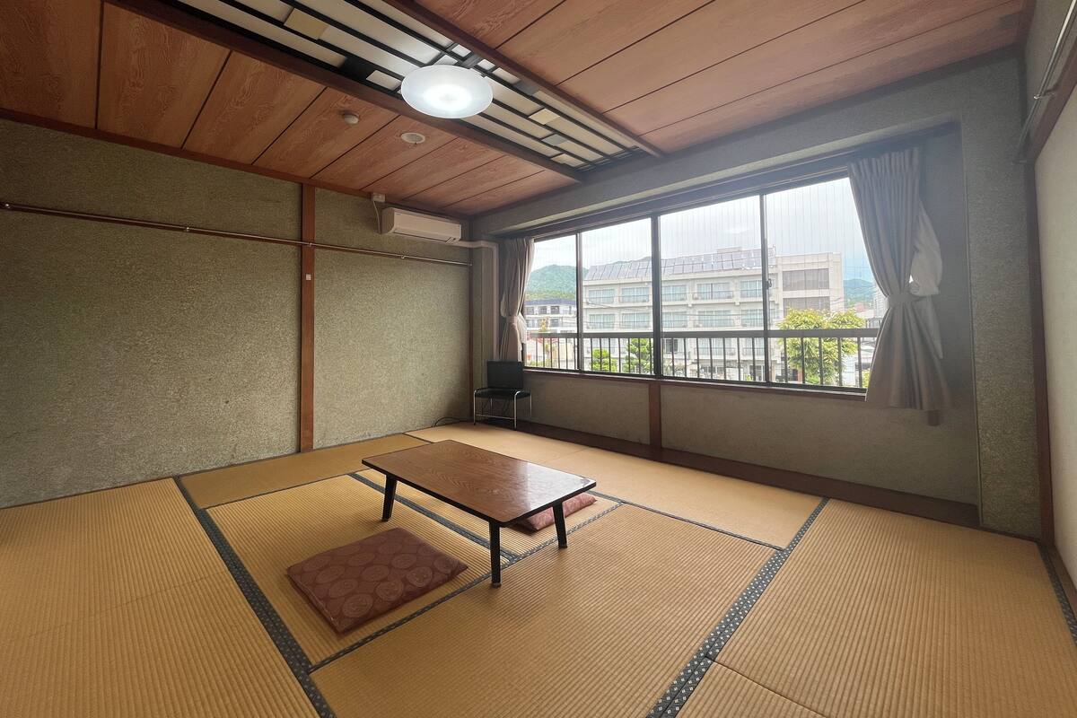 A tatami mat room with a wooden ceiling, a table, a bench, and a large window with a view of buildings.