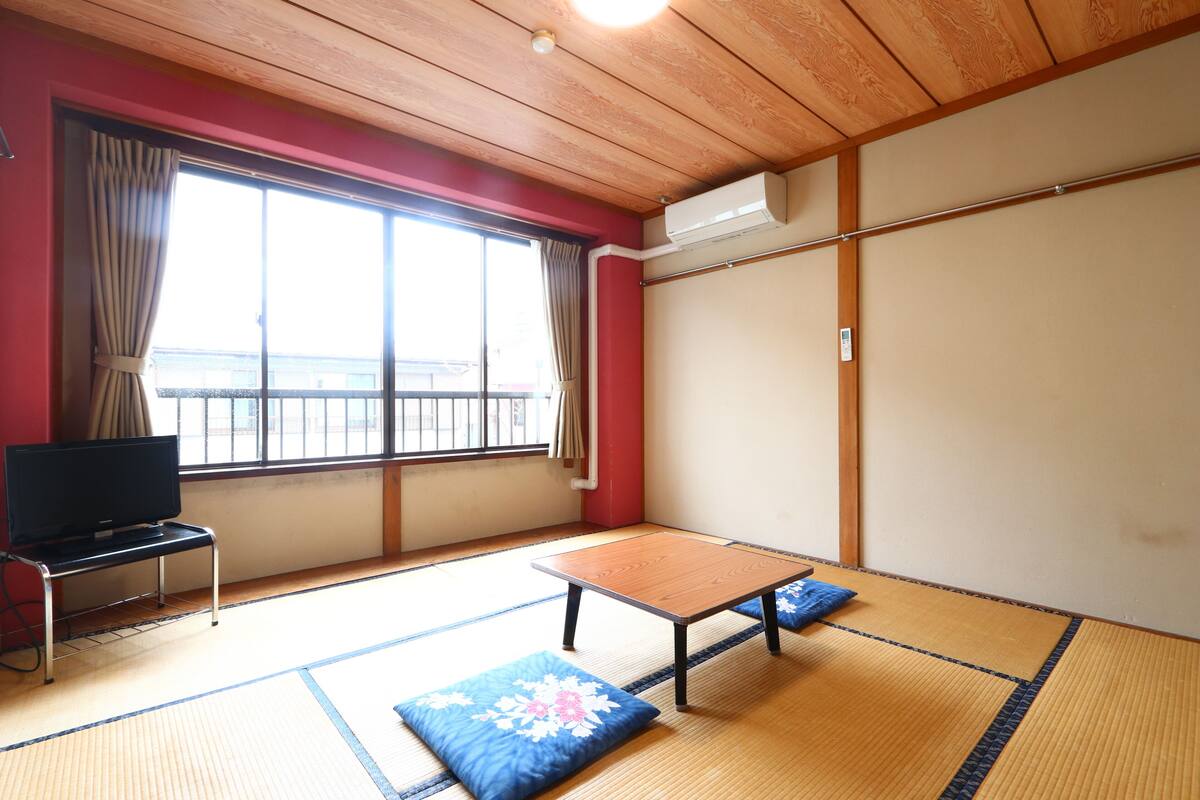 A room with tatami flooring, a television on a stand, a table, and a view of the outside through a large window.