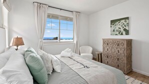 A bedroom with a bed, a window with curtains, a chair, a dresser, and a painting on the wall. - Marbella Lane - 7BR Home w Mountain Ocean Views (Waianae)