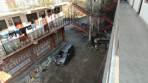 Ein mehrstöckiges Gebäude mit einem Laden im Erdgeschoss links, mit einem Schild für 'Elegant TMT Bars'. Ein silbernes Auto ist vor dem Gebäude geparkt, und Baumaterialien liegen auf dem Boden verstreut.