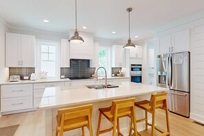A modern kitchen with white cabinets, stainless steel appliances, and a large island with bar stools. - Brand New Coastal Retreat, 5BR Dog Friendly, Cape Fear Station, Near East Beach & Shoals Club (Bald Head Island)