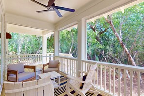 A covered porch with wooden furniture, a ceiling fan, and a view of a forest. - Brand New Coastal Retreat, 5BR Dog Friendly, Cape Fear Station, Near East Beach & Shoals Club (Bald Head Island)