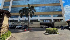 Un edificio de gran altura con fachada de vidrio moderna, un letrero que dice 'DISTRITO ARTEMISA CELAQUE' y un estacionamiento con vehículos y un guardia de seguridad.
