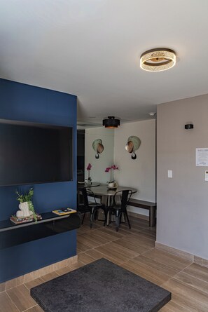 Un salon moderne doté d’un téléviseur à écran plat, d’un mur d’accent bleu foncé, d’un coin repas avec une table et des chaises, et d’un luminaire au plafond.