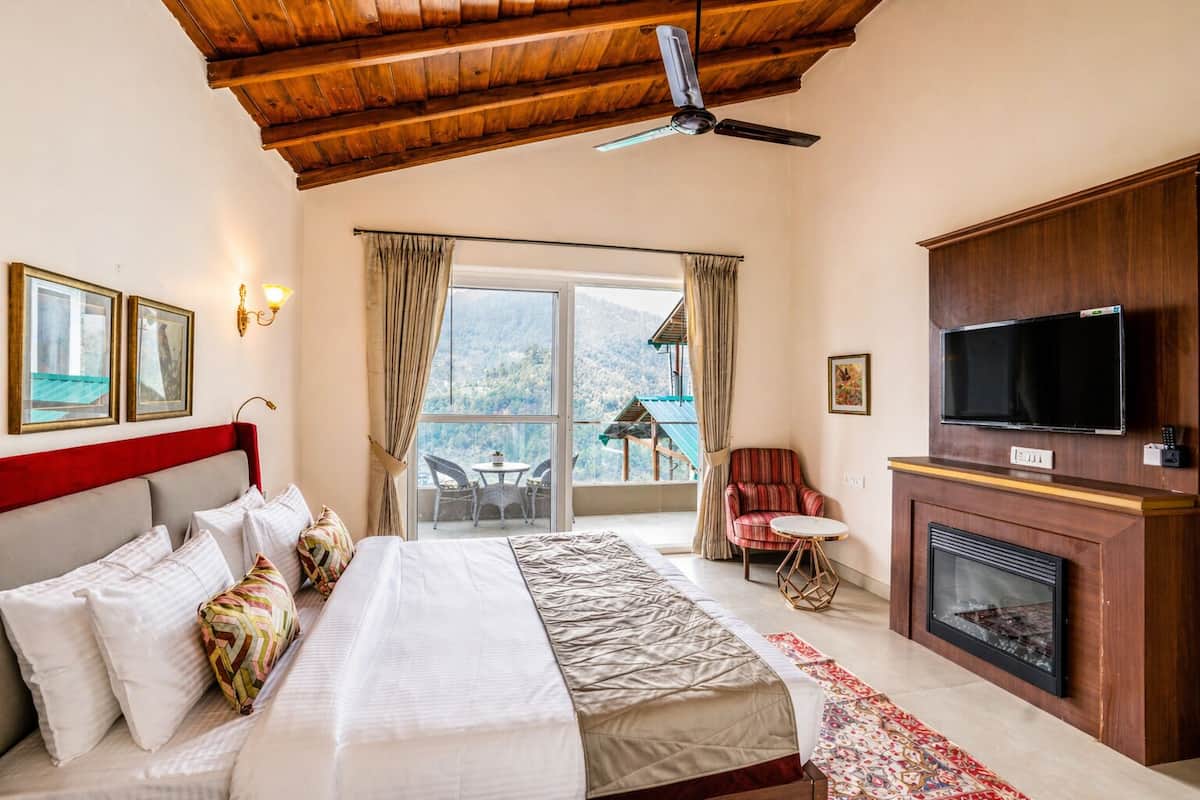 A bedroom with a bed, a TV, a fireplace, a balcony with a table and chairs, and a view of the mountains.