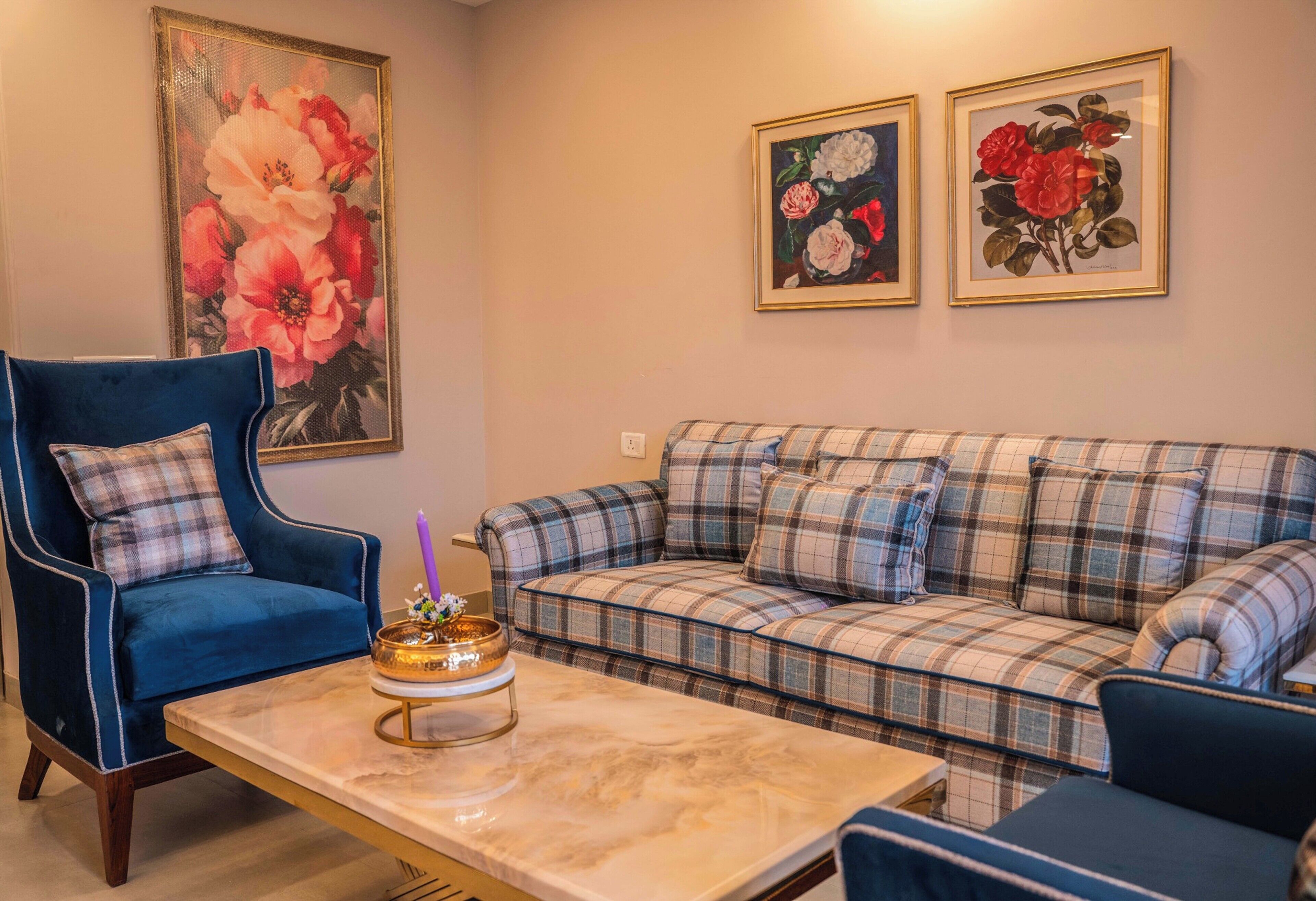 Een woonkamer met een blauwe fauteuil, een ruitjesbank, een marmeren salontafel en bloemkunstwerken in lijsten aan de muur.