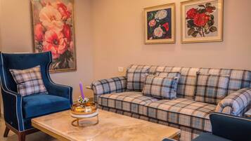 Een woonkamer met een blauwe fauteuil, een ruitjesbank, een marmeren salontafel en bloemkunstwerken in lijsten aan de muur.
