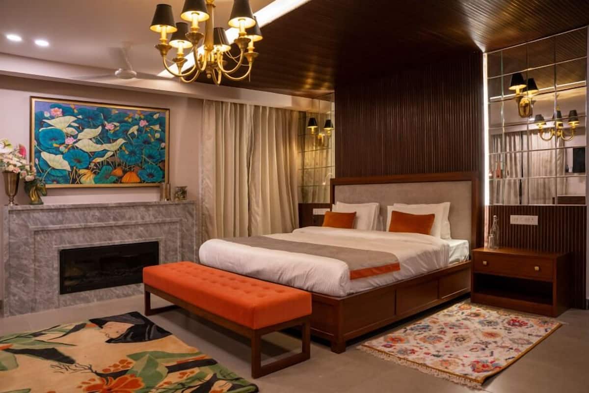 A bedroom with a large bed, a fireplace, a bench, a chandelier, and a painting on the wall.