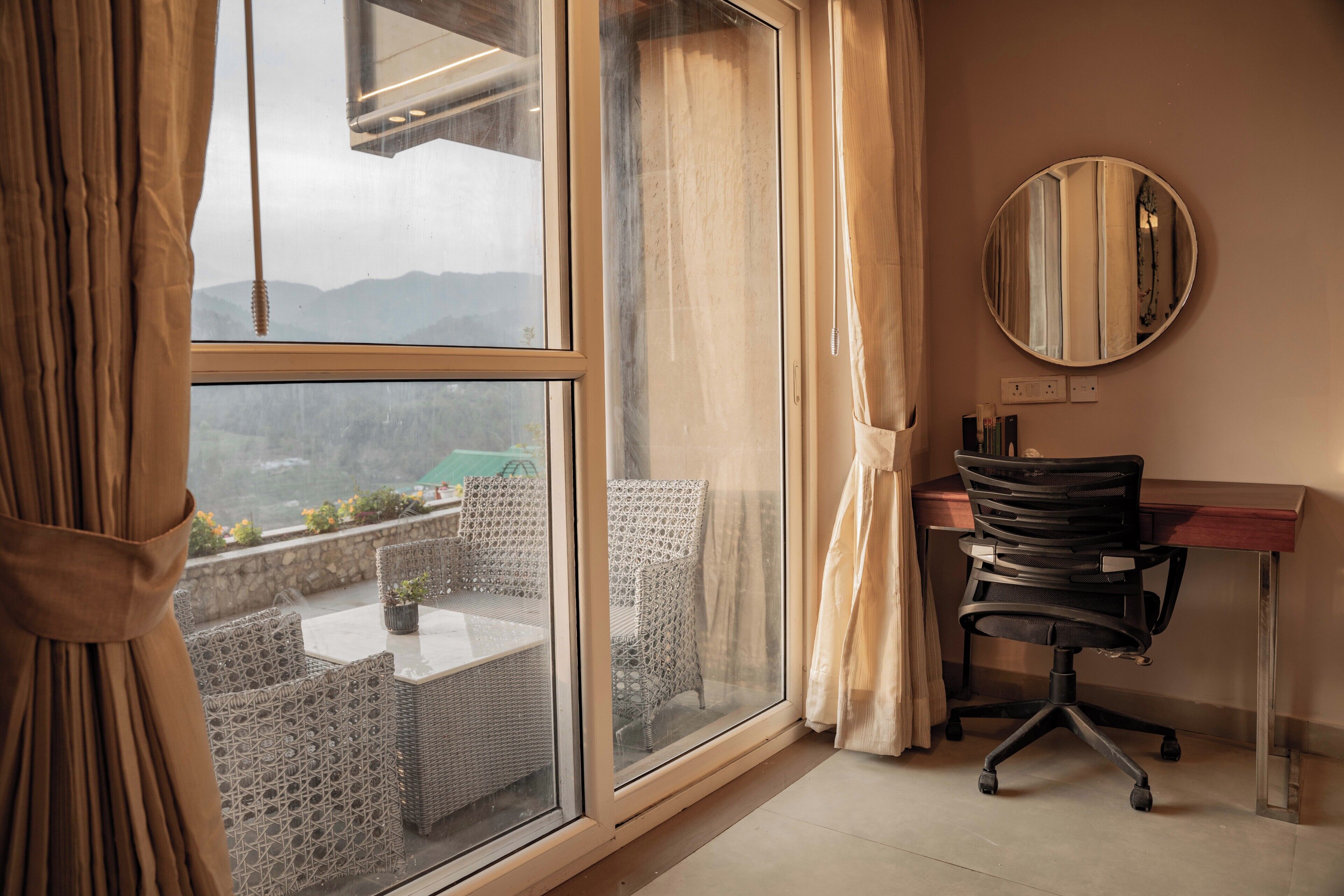 A room with a large window, a desk, a chair, and a round mirror on the wall.
