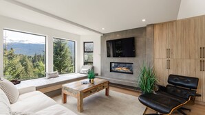 Ein modernes Wohnzimmer mit einer Couch, einem Couchtisch, einem Kamin und großen Fenstern mit Blick auf die Berge.