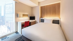 Hotellihuone, jossa on suuri sänky, työpöytä tietokoneella, punainen tuoli ja näkymä kaupunkiin.