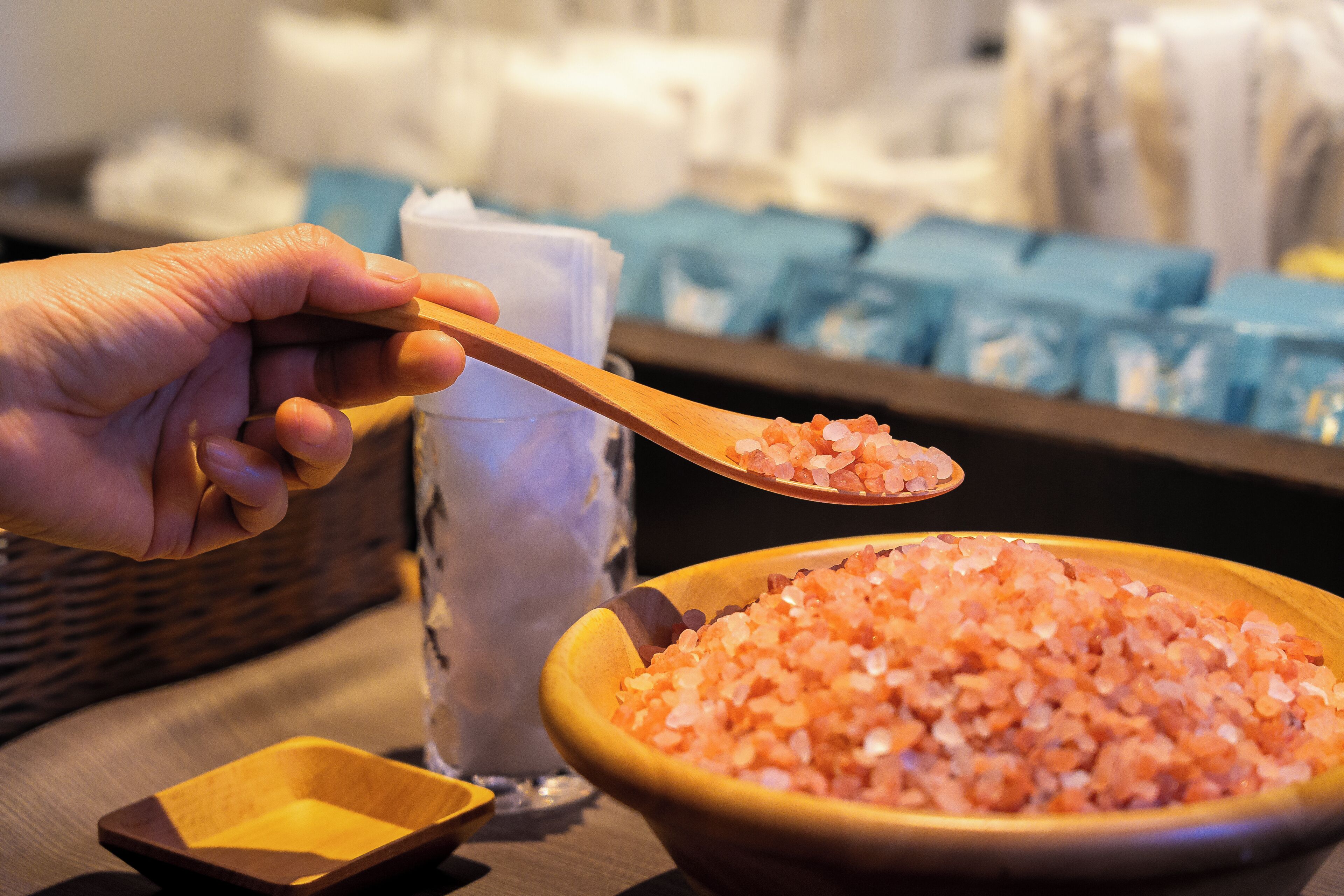 A hand holding a wooden spoon over a bowl of pink salt crystals, with a small wooden bowl and a glass container in the background.