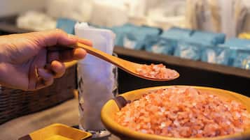 A hand holding a wooden spoon over a bowl of pink salt crystals, with a small wooden bowl and a glass container in the background.