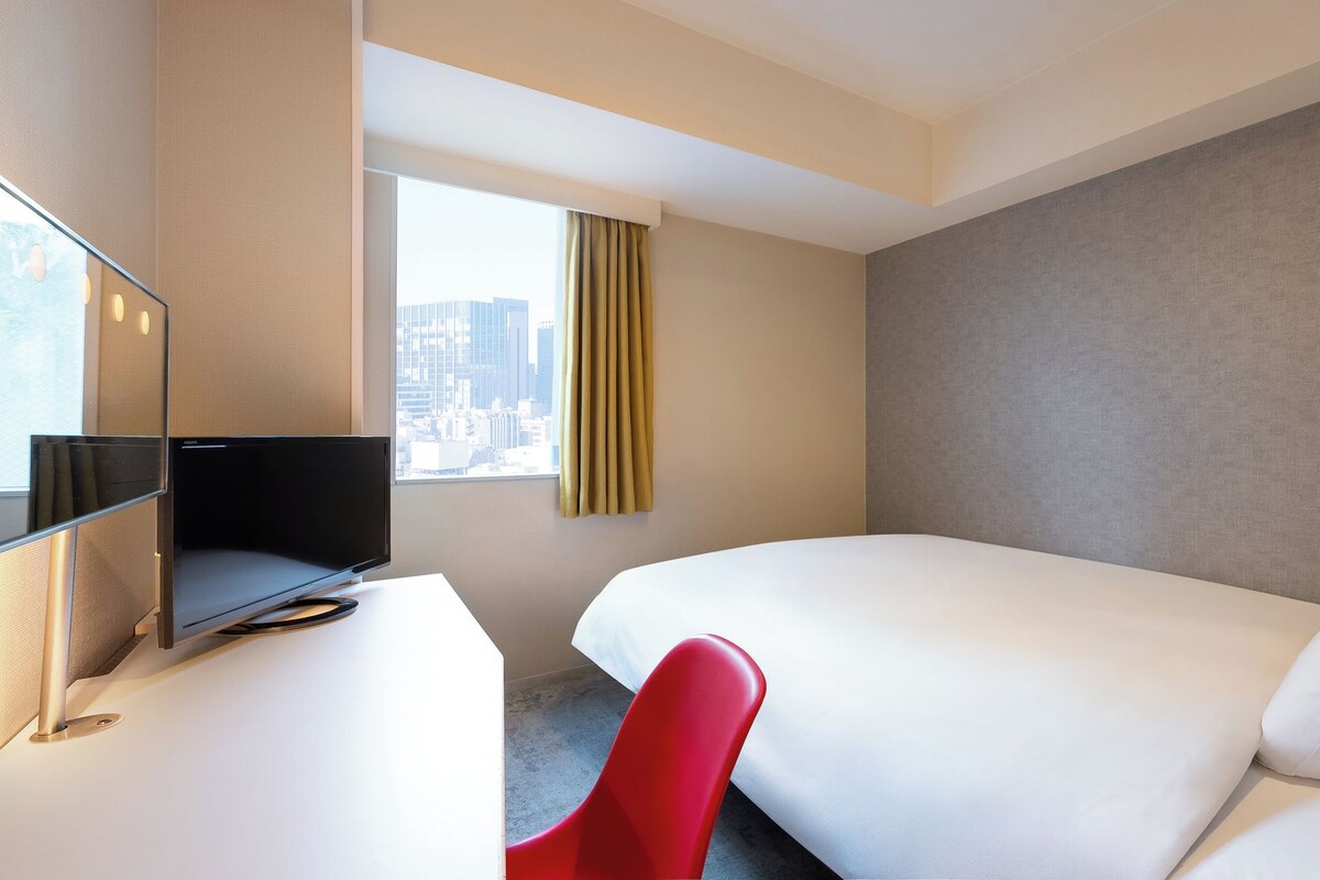 A hotel room with a bed, a desk with a computer, a red chair, and a window with curtains.