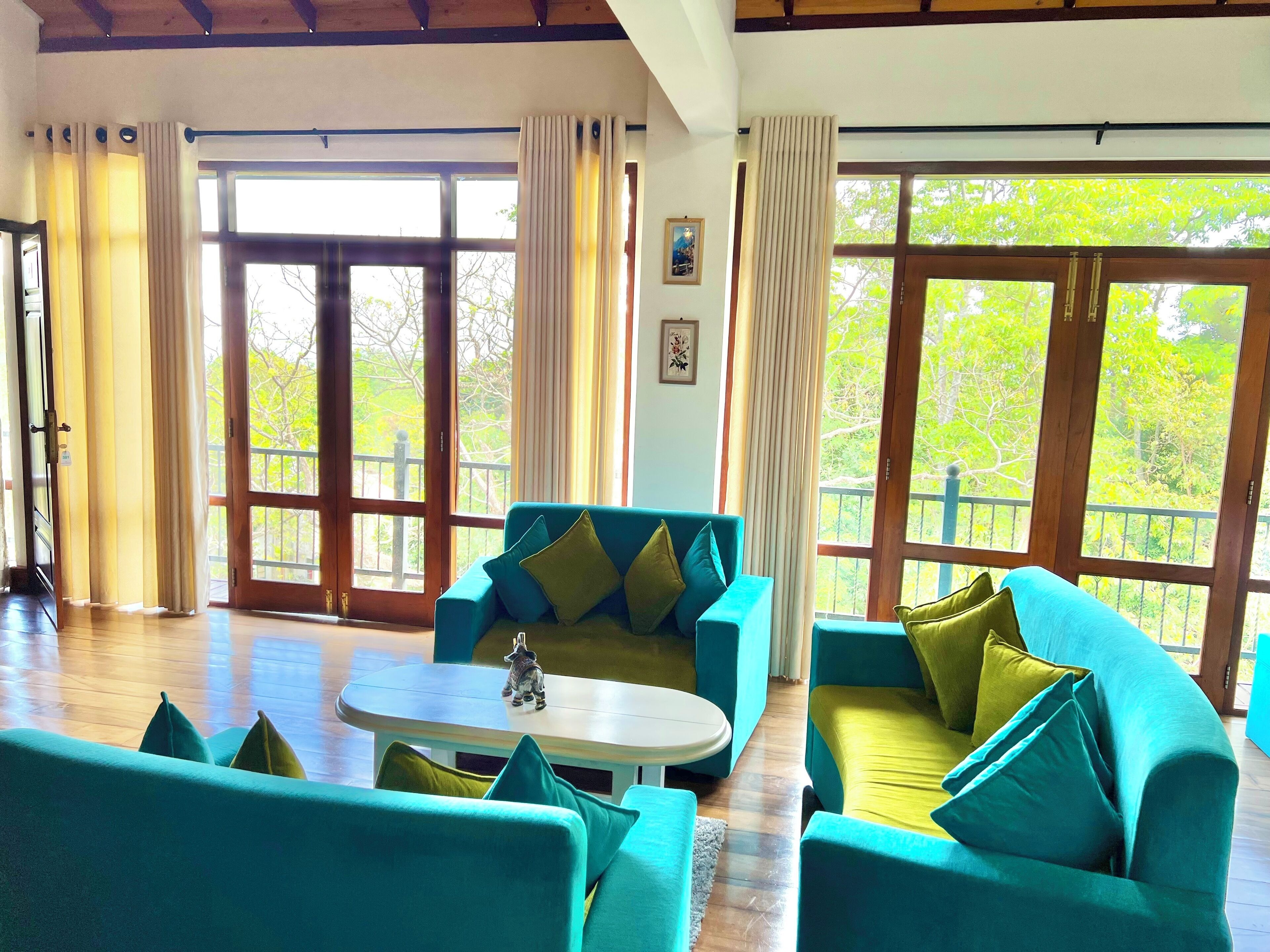 Amplia sala de estar con sofás color turquesa y una mesa de centro blanca, ventanas grandes con cortinas y piso de madera.