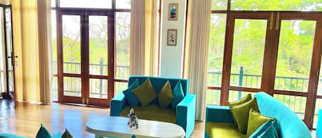 Amplia sala de estar con sofás color turquesa y una mesa de centro blanca, ventanas grandes con cortinas y piso de madera.