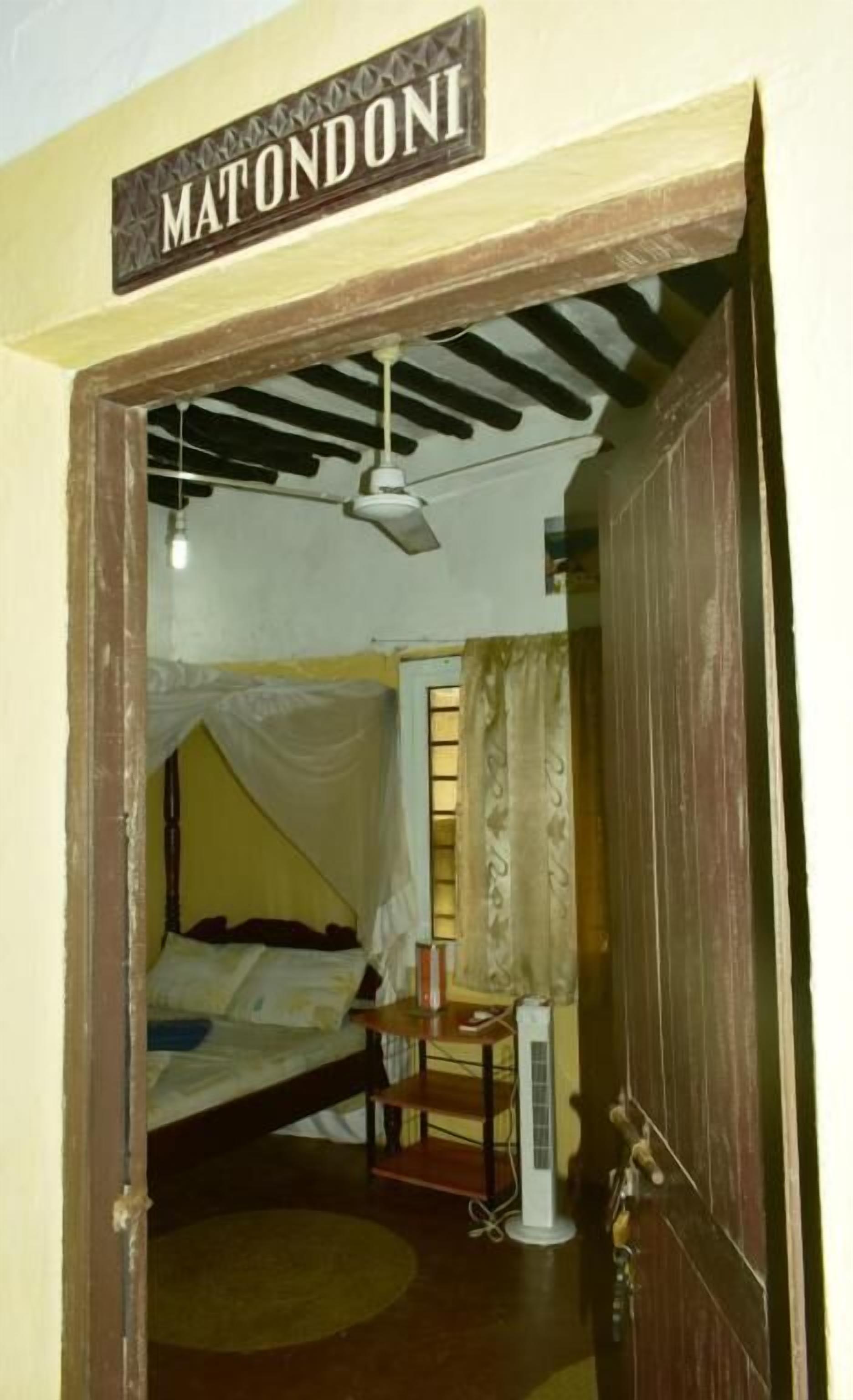 Una habitación con una cama, un ventilador de techo, un calentador y una puerta con la leyenda "MATONDONI".