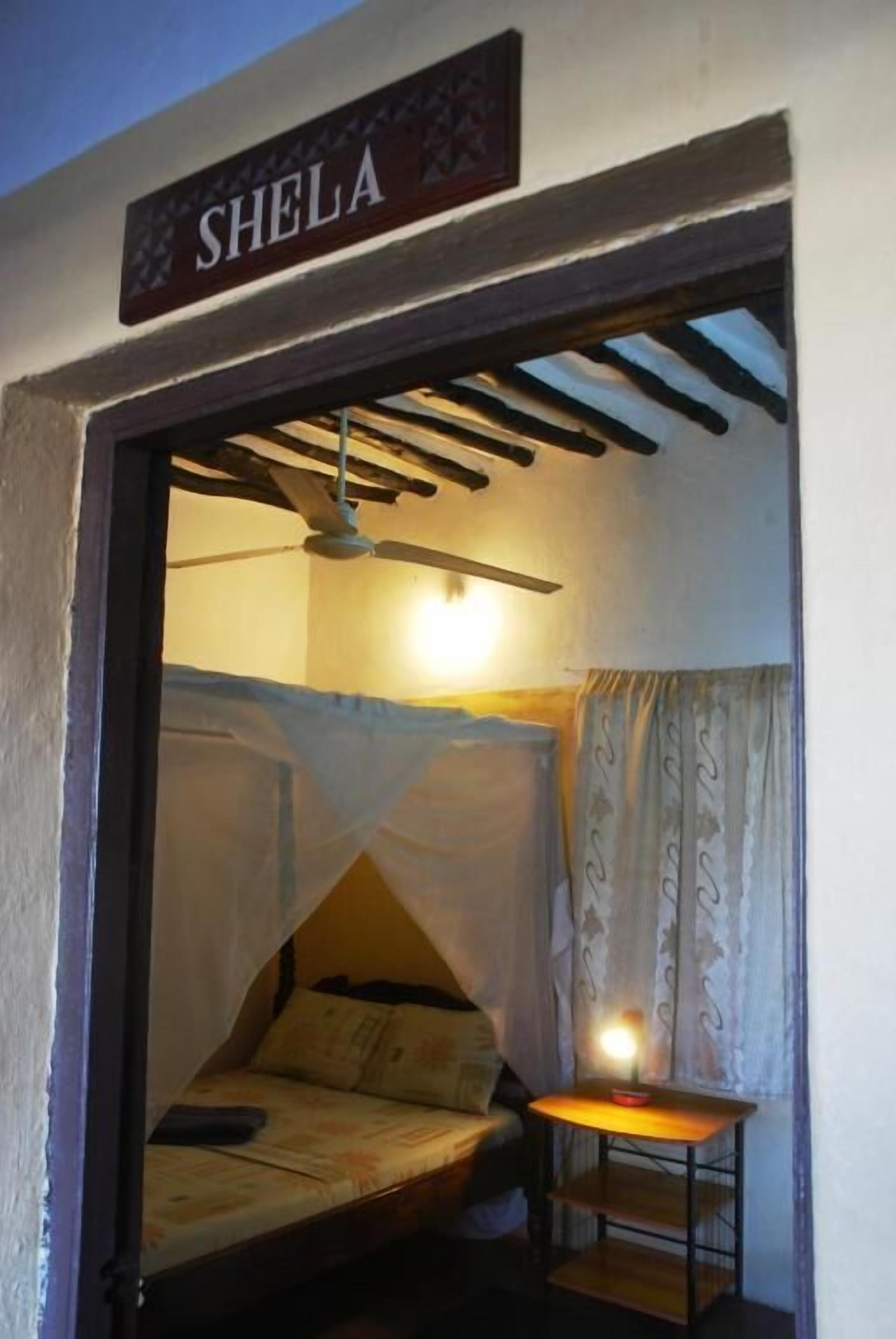 A room with a bed, a ceiling fan, a table with a lamp, and a sign that reads "SHELA".