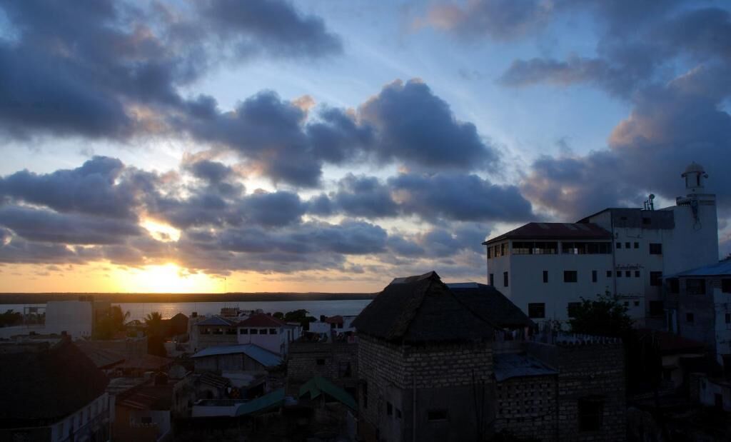 A coastal town with a prominent white building with a tower, surrounded by stone structures and a sunset in the background.