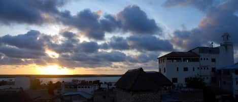 A coastal town with a prominent white building with a tower, surrounded by stone structures and a sunset in the background.