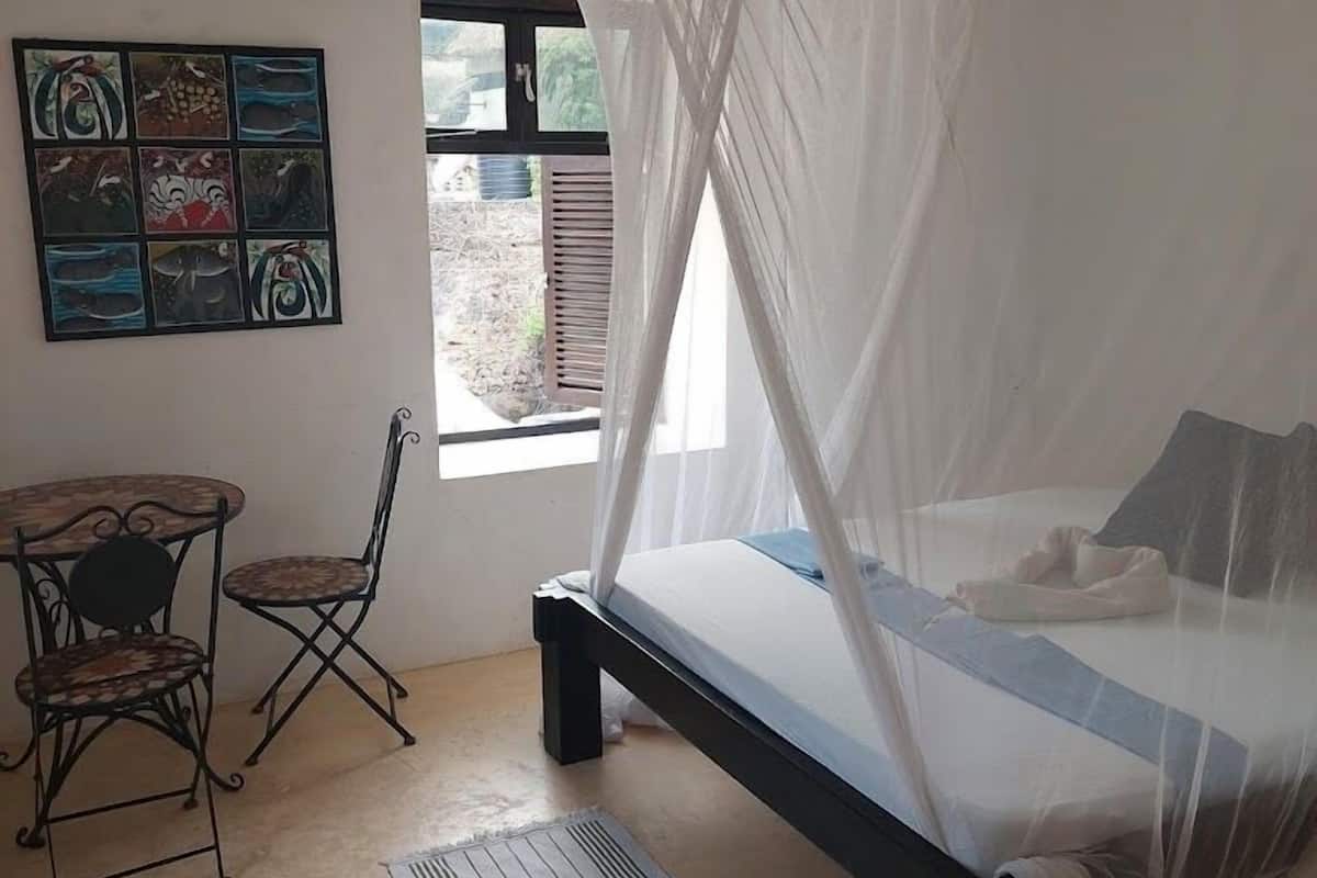 A bedroom with a bed under a canopy, a small table with chairs, and a window with a view of the outside.