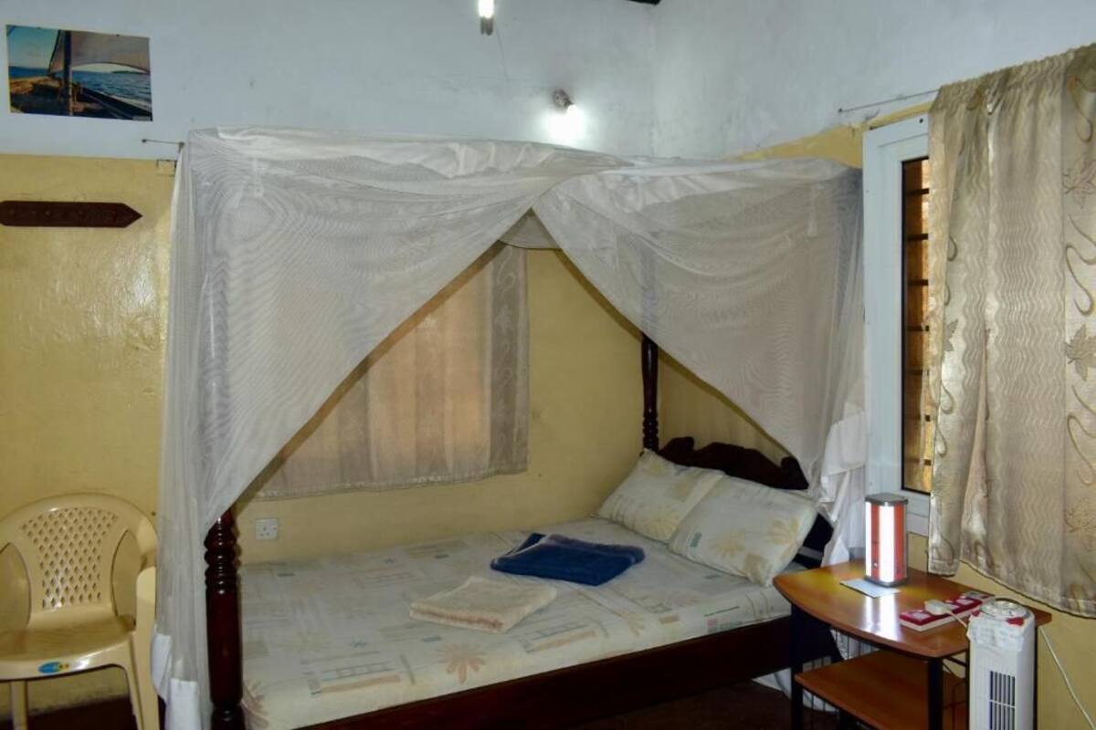 A single bed with a canopy, a bedside table with a lamp, a chair, and a window with curtains.