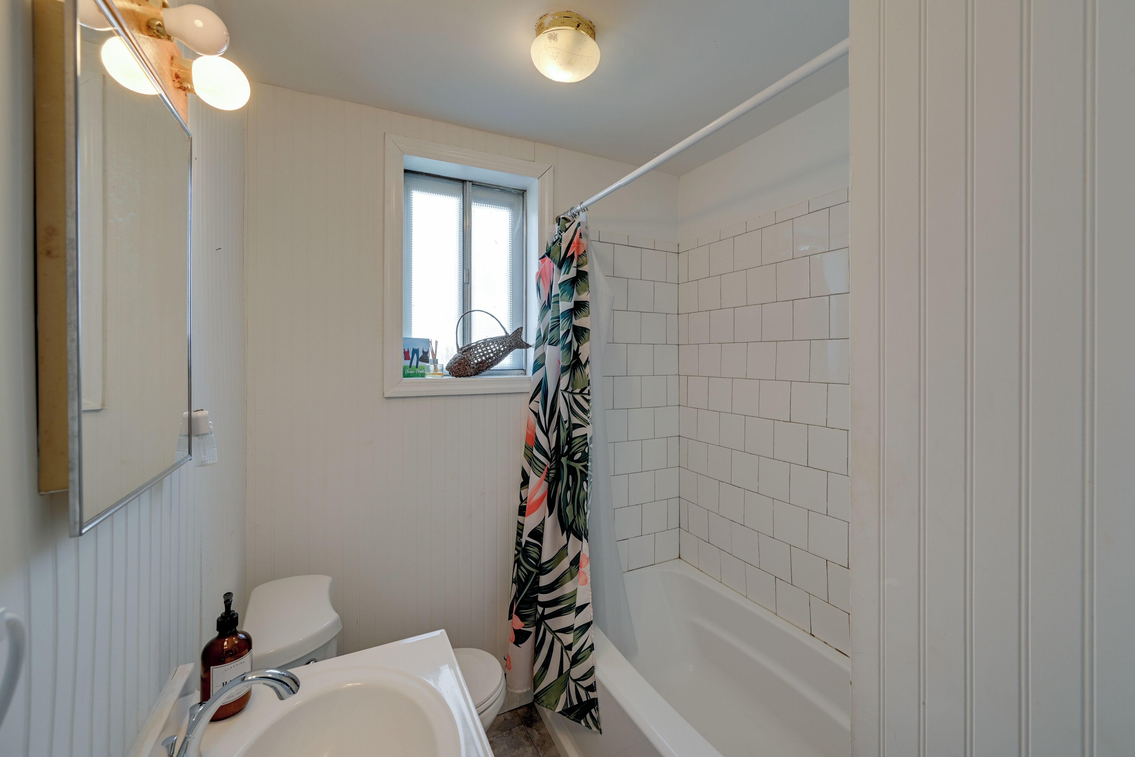 Une salle de bain avec des toilettes blanches, un lavabo blanc avec un robinet, une douche avec un rideau aux motifs de feuilles colorées et une fenêtre au-dessus des toilettes.