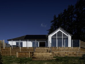 A modern building with a sloped roof, large windows, and a wooden deck leading to the entrance. - Sunny Ridge is a modern, open-plan, sustainable bungalow, with stunning views. (Fort Augustus)