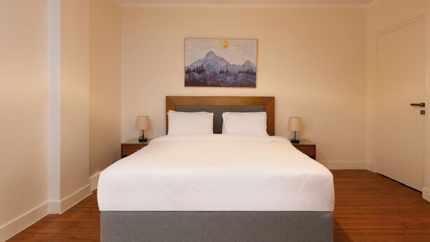 A neatly made bed with a wooden headboard, two bedside tables with lamps, and a painting of a mountain landscape on the wall. - The Heart of Heliopolis 76 El Hegaz St (Cairo)