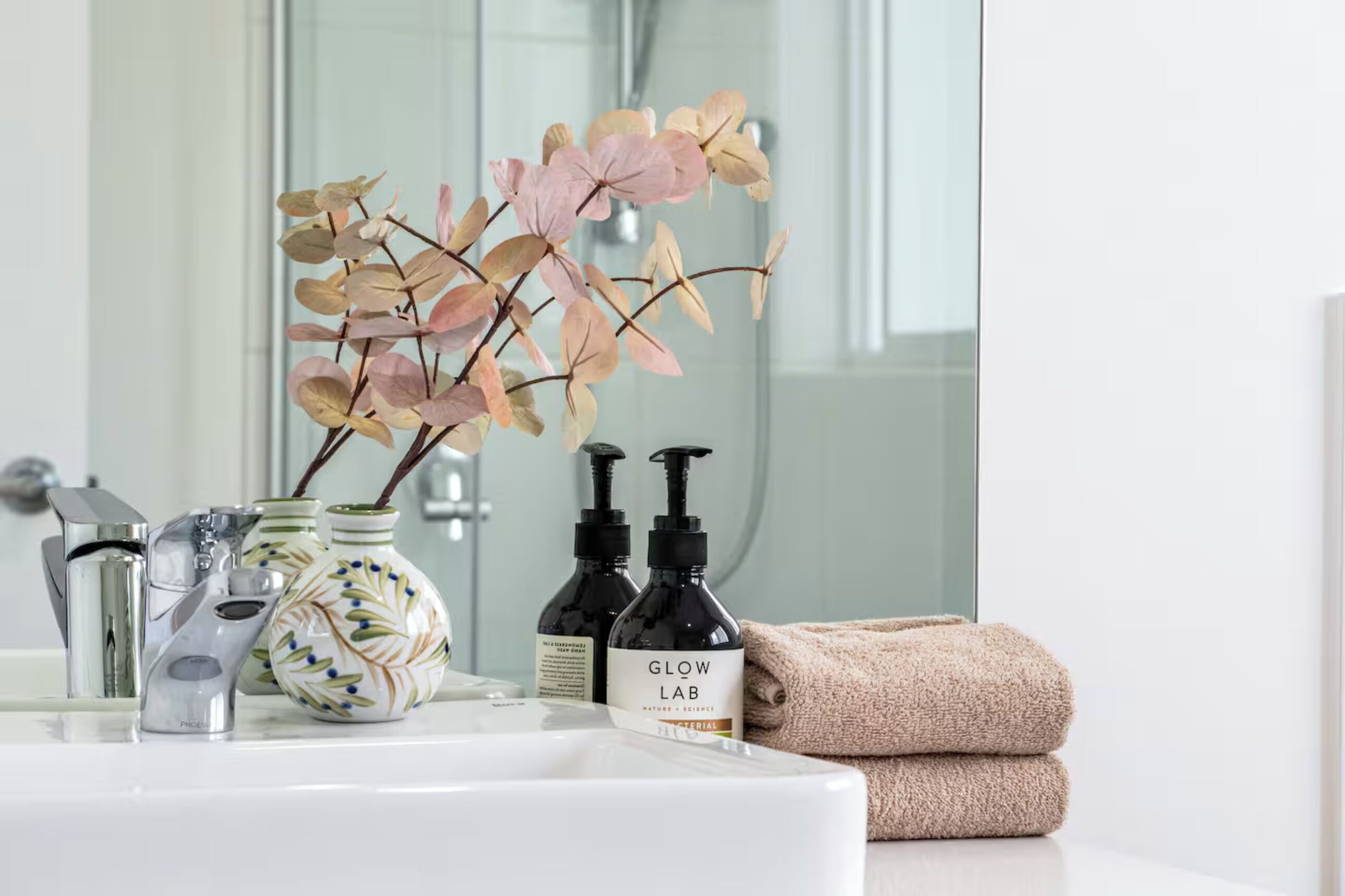 A bathroom vanity with a white sink, a decorative vase with dried flowers, two black bottles labeled "GLOW LAB," and a folded beige towel.