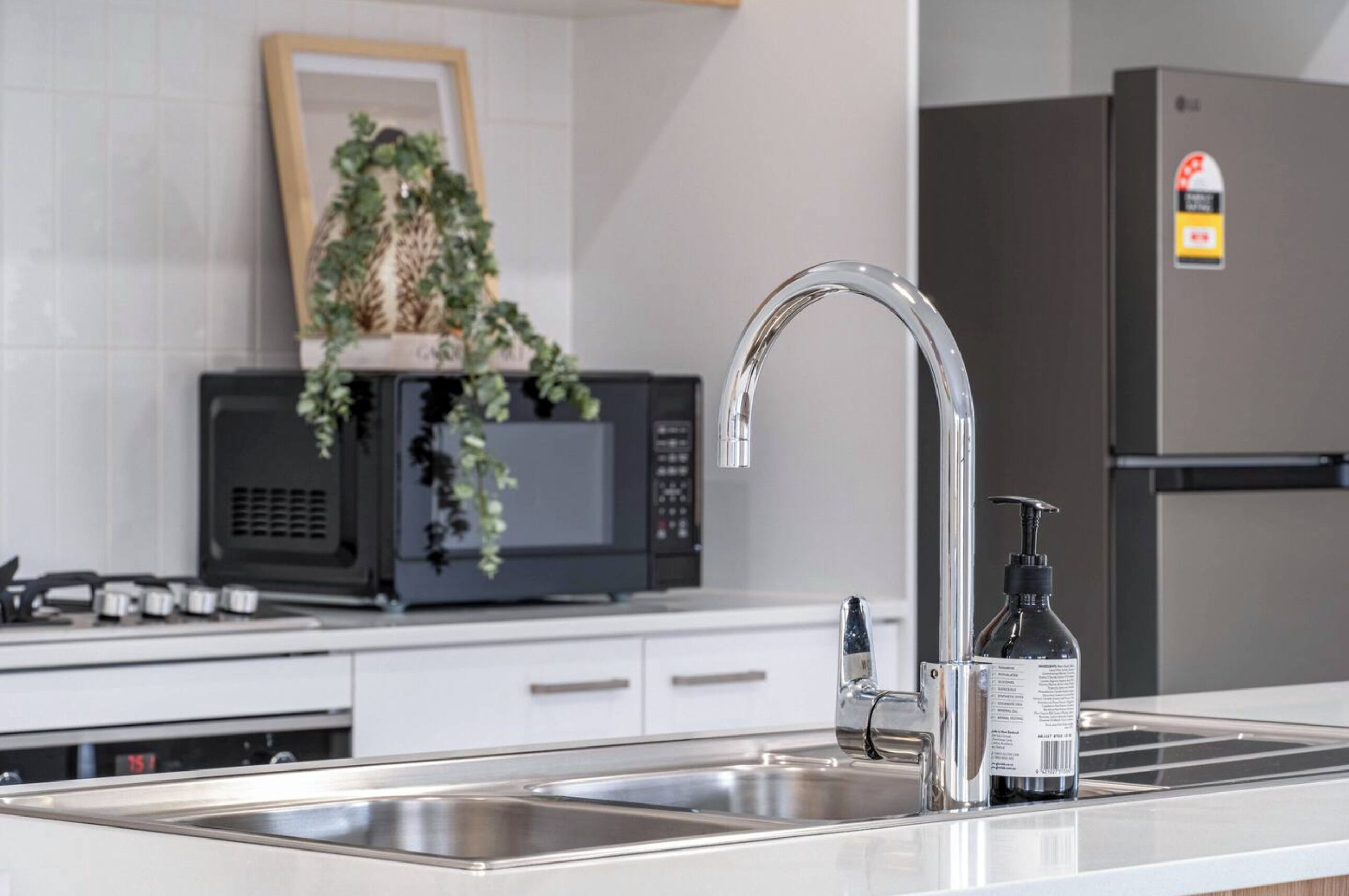Une cuisine moderne dotée d’un évier en acier inoxydable, d’un micro-ondes noir, d’un réfrigérateur et d’une plante décorative posée sur le micro-ondes.