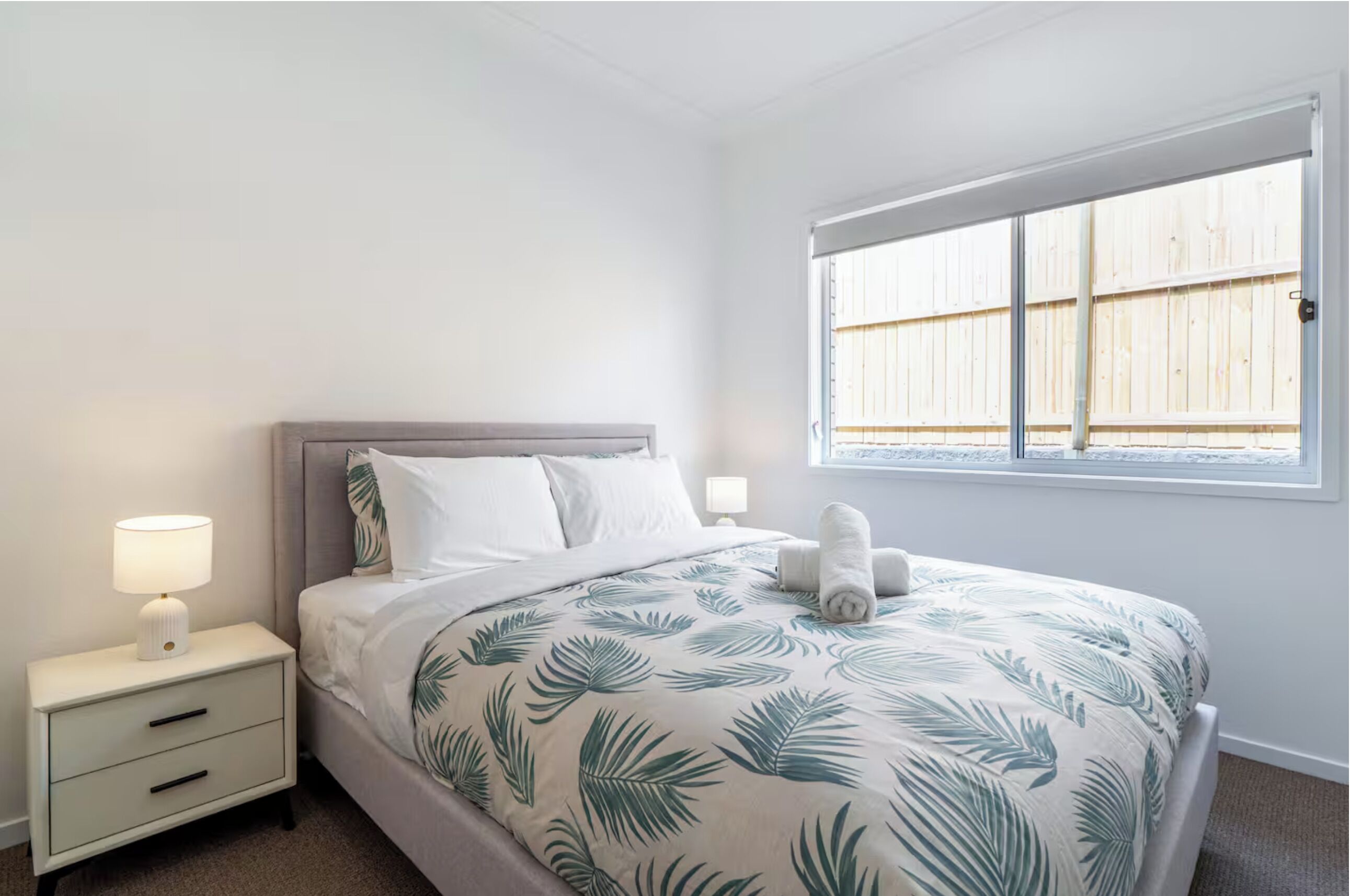 A bedroom with a bed, nightstands, and a window with a view of a wooden fence.