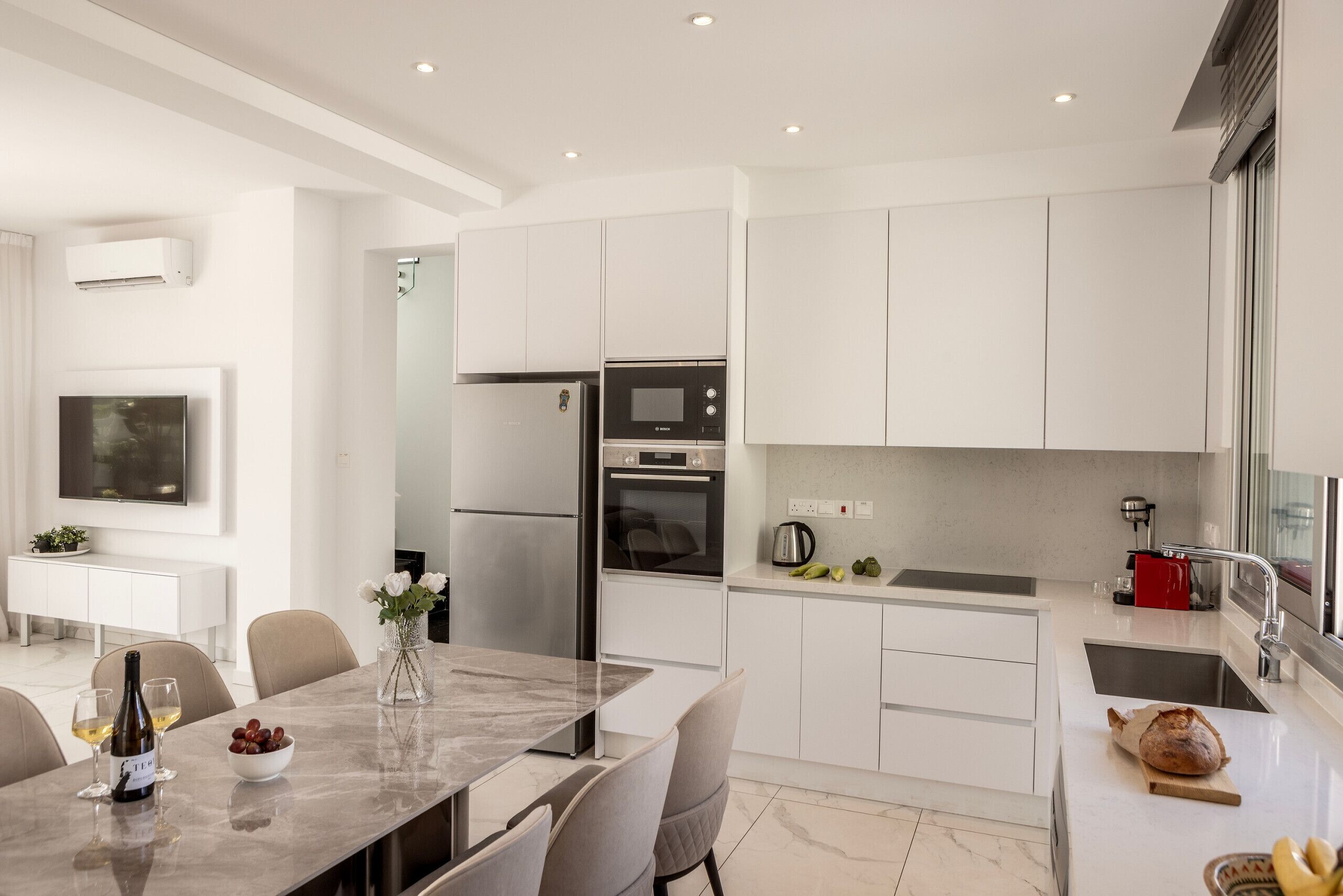 A modern kitchen with white cabinets, a stainless steel refrigerator, an oven, and a microwave. There is a dining table with chairs, a vase with flowers, and a bottle of wine.