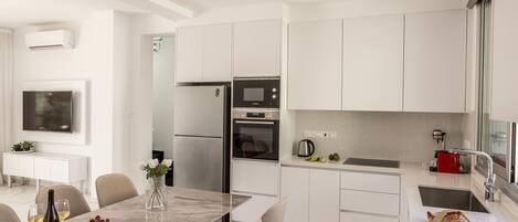 A modern kitchen with white cabinets, a stainless steel refrigerator, an oven, and a microwave. There is a dining table with chairs, a vase with flowers, and a bottle of wine.