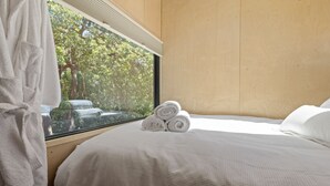 Quarto com uma grande janela, paredes de madeira, cama bem arrumada com toalhas dobradas e roupão branco pendurado perto da janela.