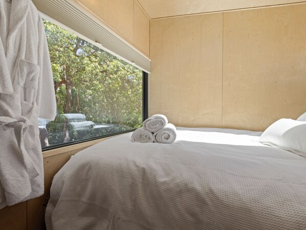 Un dormitorio con un ventanal grande, paredes de madera, una cama bien tendida con toallas enrolladas y una bata blanca colgada junto a la ventana.
