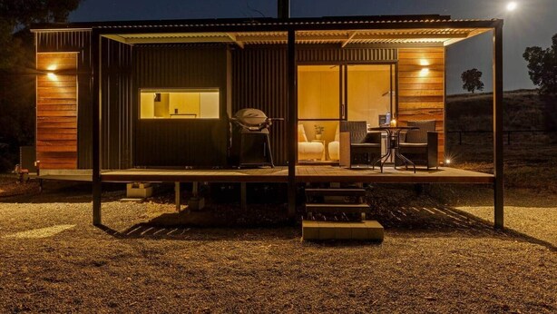 Une cabane moderne dotée d’un porche couvert, d’une grande fenêtre et d’un espace salon avec des chaises et une table.