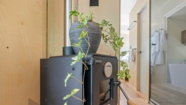 A compact wood-burning stove with a large vase and a potted plant on top, situated in a hallway with a wooden floor and a glass door leading to a bathroom. - Off Grid Tiny Home Fern View: Escape the City, Unwind & Reconnect with Nature (North Dandalup)