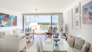 Ein geräumiger Wohnbereich mit einem weißen Sofa, einem Esstisch mit Stühlen und Meerblick durch große Fenster.
