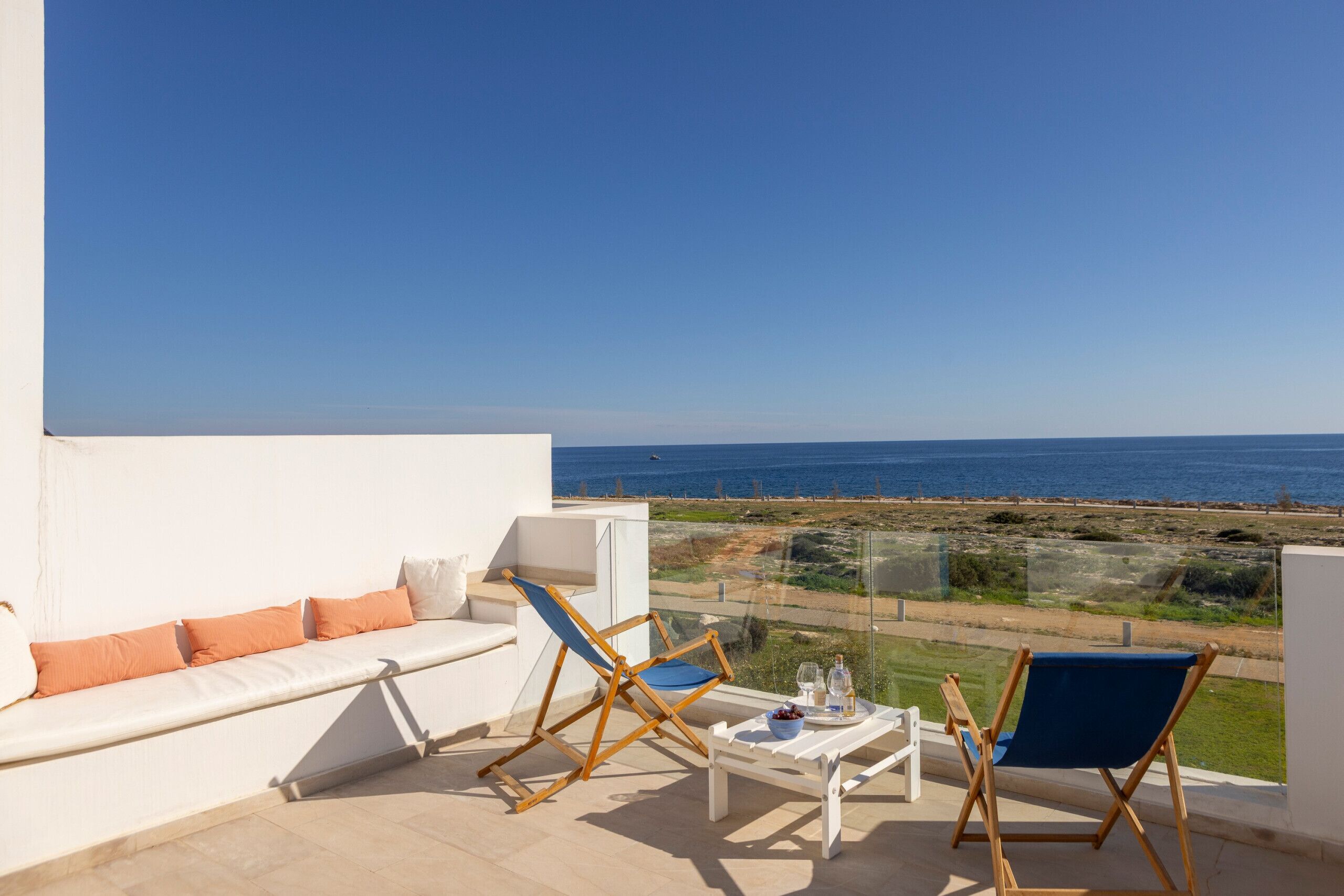 A balcony with a white sofa, two folding chairs, a small table with a tray, and a clear view of the sea and beach.