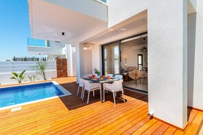 A modern outdoor patio with a pool, wooden deck, and a dining area with white chairs and a table set with fruits and a vase. - Protaras Sol e Mar Villa 1 (Paralimni)