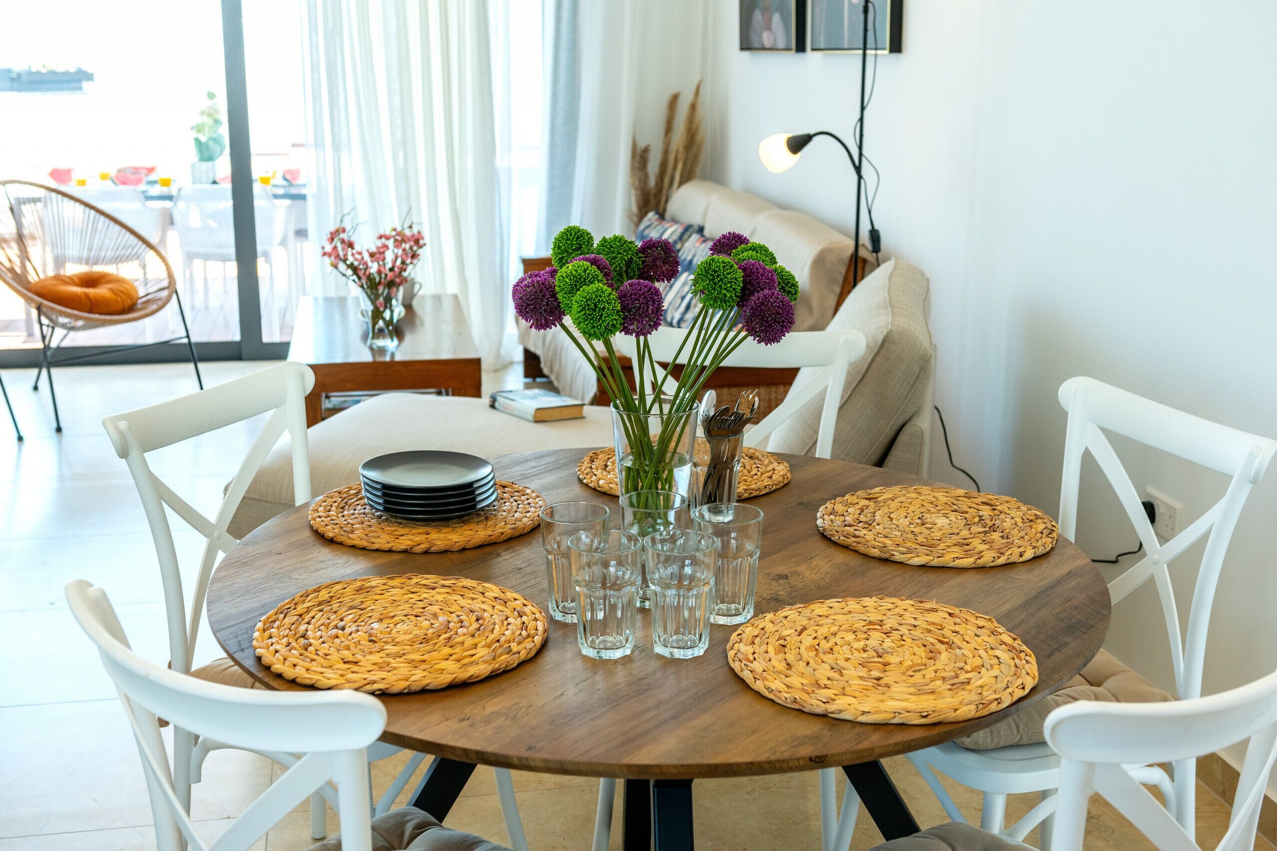 A dining table with woven placemats, clear glasses, and a vase with purple flowers.