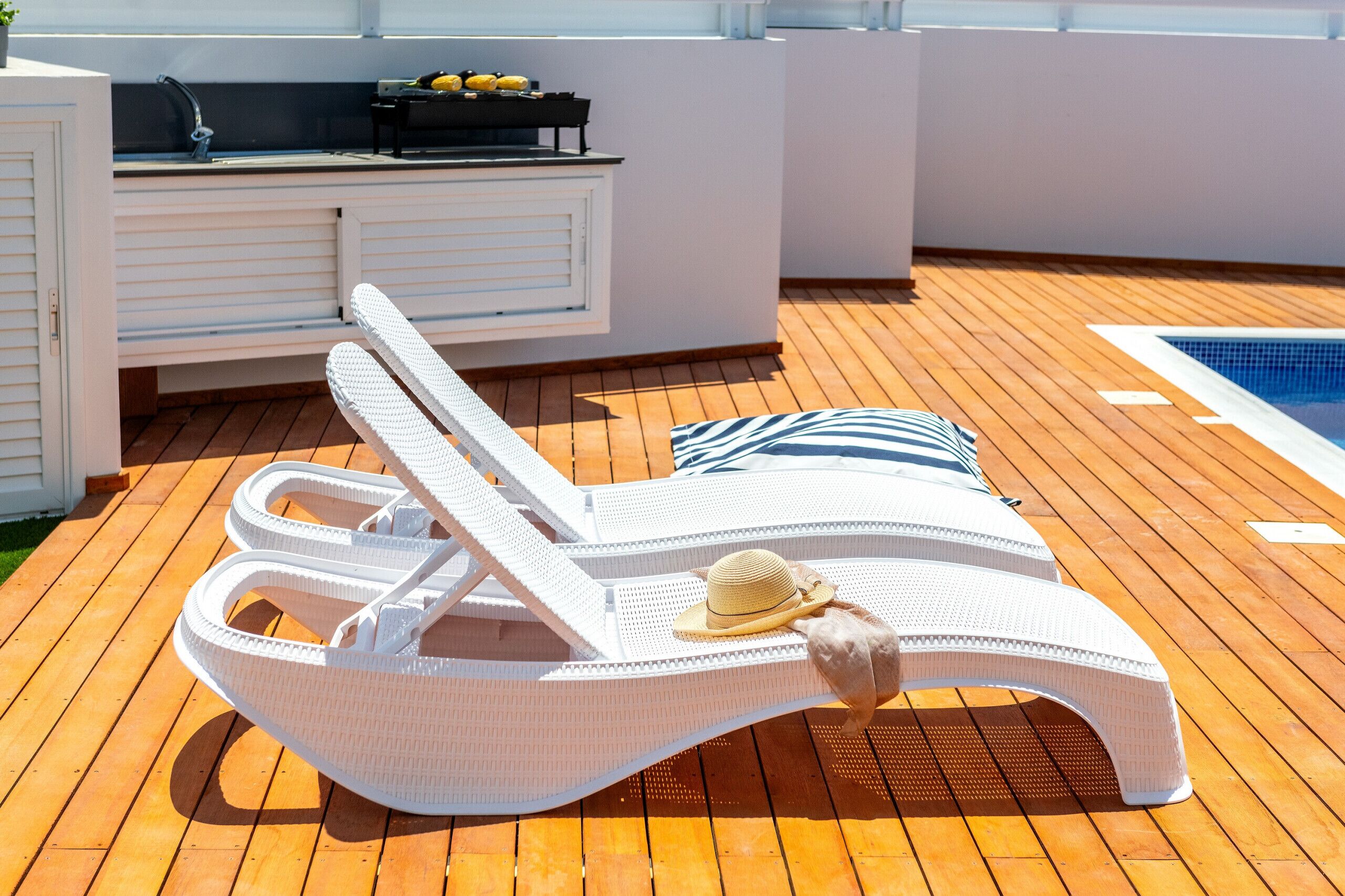 A sun lounger with a hat, a pool, and a barbecue area on a wooden deck.
