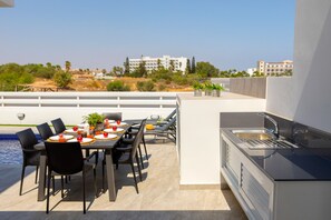 Una terrazza sul tetto con un tavolo da pranzo per quattro persone, una cucina integrata con lavello e vista su edifici e vegetazione sullo sfondo.