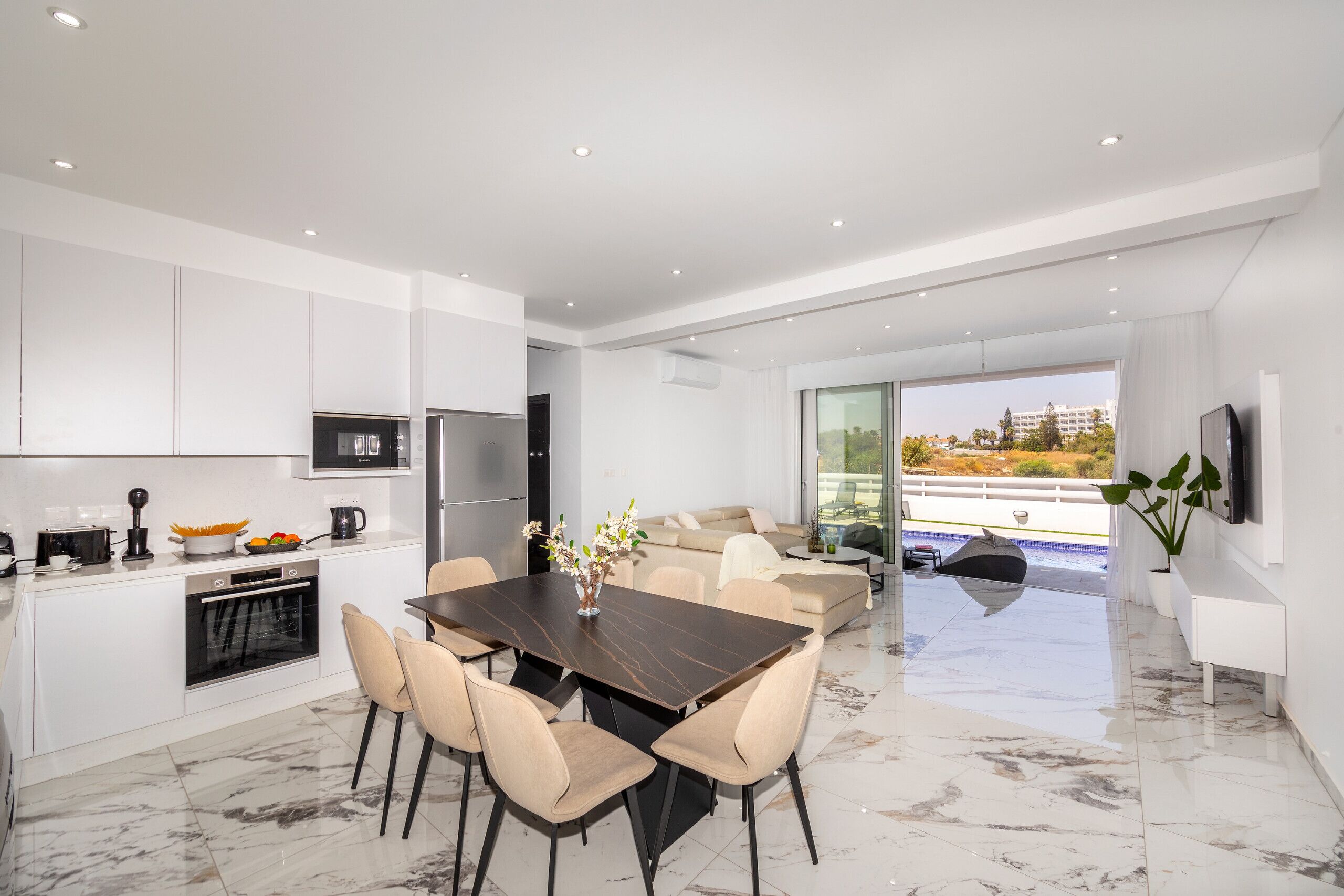 Una cocina moderna con armarios blancos, un comedor con mesa de mármol y una sala de estar con sofá y un amplio ventanal.
